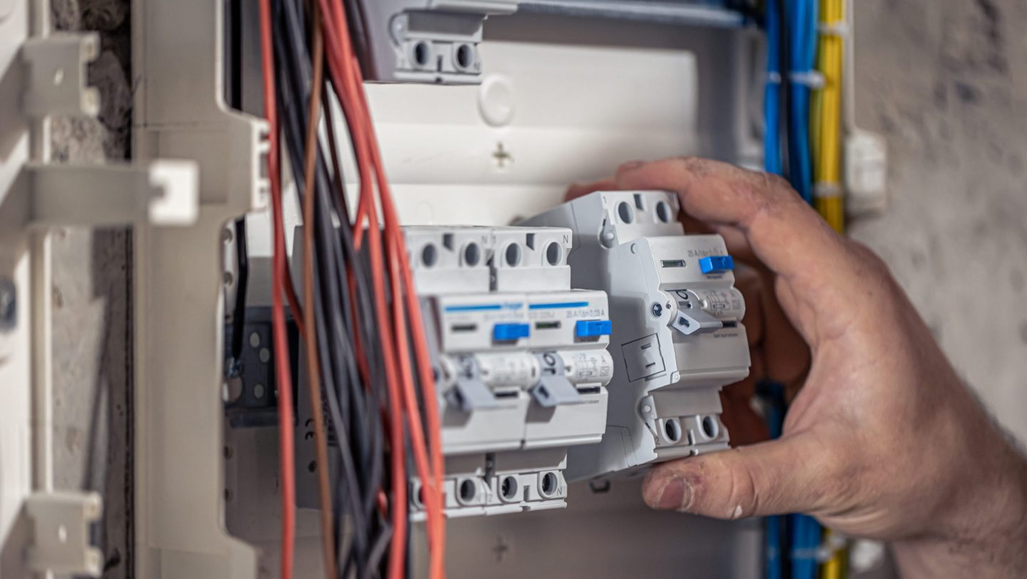 A male electrician works in a switchboard with an electrical connecting cable, connects the equipment with tools.
